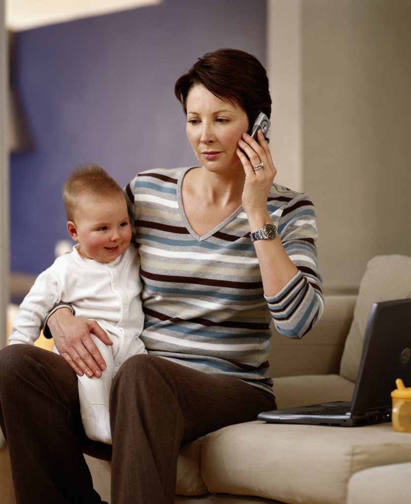 Πολυάσχολη μητέρα χωρίς ελεύθερο χρόνο για τον εαυτό της που κάθεται στον καναπέ κρατώντας ένα παιδί στα χέρια της ενώ ταυτόχρονα είναι απασχολημένη μιλώντας στο τηλέφωνο